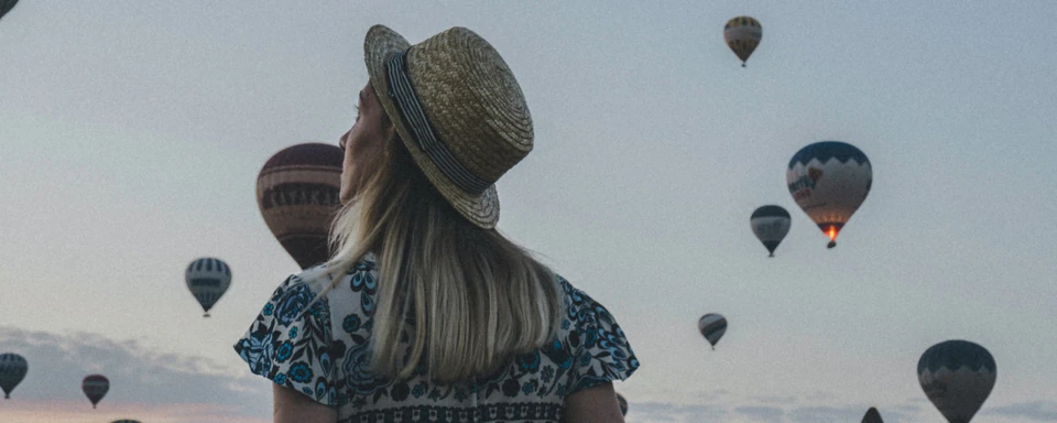 A traveler in a straw hat watching hot air balloons fill the dawn sky over Cappadocia.