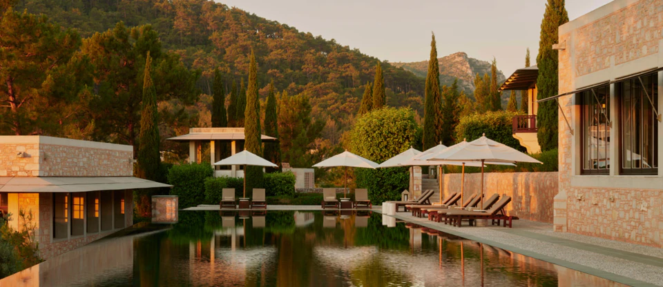 A resort pool lined with sun loungers, umbrellas, and cypress trees at golden hour.