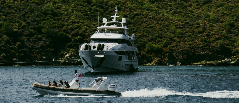 A motoryacht at anchor as a yacht club tender speeds past in a green-hilled bay.