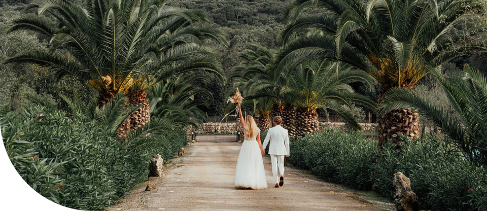 Newlyweds walking hand in hand down a palm-lined lane, bouquet raised