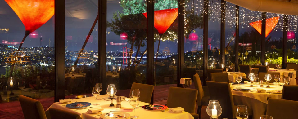 Candlelit tables at a hillside restaurant overlooking the Bosphorus and its illuminated bridge at night.