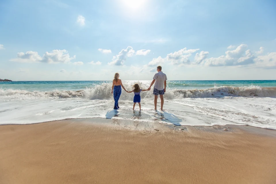 Parents and their child holding hands at the water’s edge on a sunlit beach.
