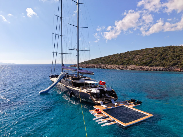 A dark-hulled two-masted sailing yacht at anchor in a turquoise cove